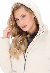 Blonde vrouw met krullend haar draagt een crèmekleurige pufferjack met capuchon over een blauw en bruin geruit overhemd, terwijl ze haar haar bij haar gezicht aanraakt.