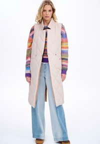 Beżowa pikowana długa kamizelka z kołnierzem, napami oraz bocznymi kieszeniami, nałożona na wielokolorowy sweter w paski i spodnie z szerokimi nogawkami.