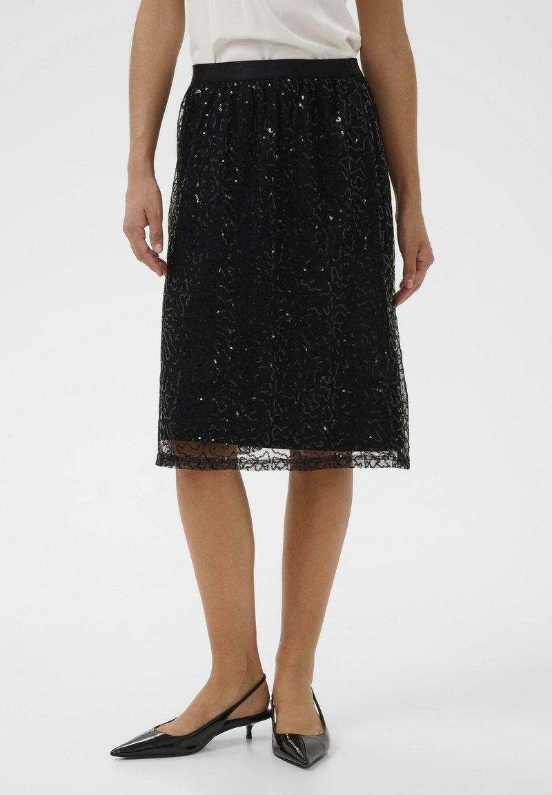 Falda negra hasta la rodilla con una superposición de encaje texturizado y patrones intrincados, cinturilla elástica y dobladillo transparente. Combinada con tacones negros tipo slingback.