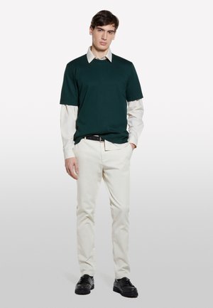 T-shirt à manches courtes vert foncé sur une chemise à manches longues rayée claire, associé à un pantalon blanc et des chaussures noires, debout contre un fond uni.