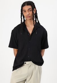 Camicia nera a maniche corte con bottoni, caratterizzata da un motivo testurizzato, con colletto classico e bottoni frontali, abbinata a pantaloni beige chiaro.