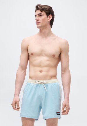 Ung mann uten skjorte iført lyseblå shorts med snøring, stående med avslappede hender, ser til venstre mot en ensfarget bakgrunn.