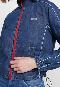 Jeansjacka i mörkblått med röd dragkedja och vita sömmar, med ståkrage och elastiska ärmslut, samt sidofickor.