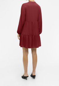 Robe fluide bordeaux avec manches longues, ourlet à volants et encolure arrondie. Portée avec des talons ouverts noirs. Texture lisse.