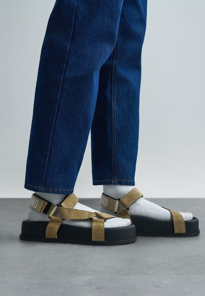 Osoba v modrých džínách, bílých ponožkách a béžových semišových sandálech s černými platformovými podrážkami, stojící na šedé betonové podlaze.