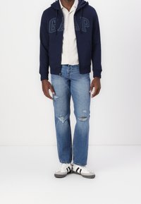 Sweat à capuche zippé bleu marine avec logo "GAP" en lettres contrastées, chemise claire en dessous, jean bleu délavé et baskets blanches à rayures noires.