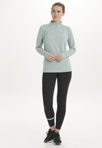 Ljusgrön, långärmad, dragkedja idrottströja i kombination med svarta leggings. Funktioner inkluderar slät tyg och en reflekterande detalj nära ankeln.