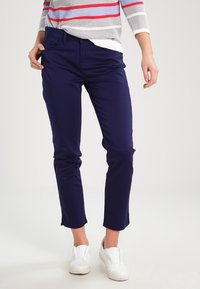 Žena nosí námořnické modré slim fit kalhoty, bílé tenisky a zkrácený pruhovaný svetr s červenými, šedými a bílými horizontálními pruhy.