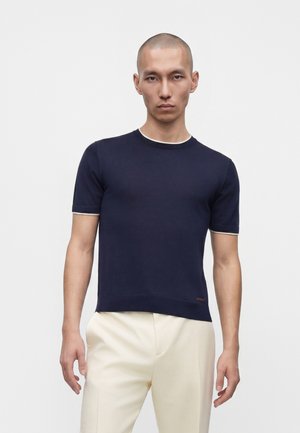 Homme au crâne rasé portant une chemise ajustée bleu marine à manches courtes et un pantalon beige clair sur fond blanc uni.