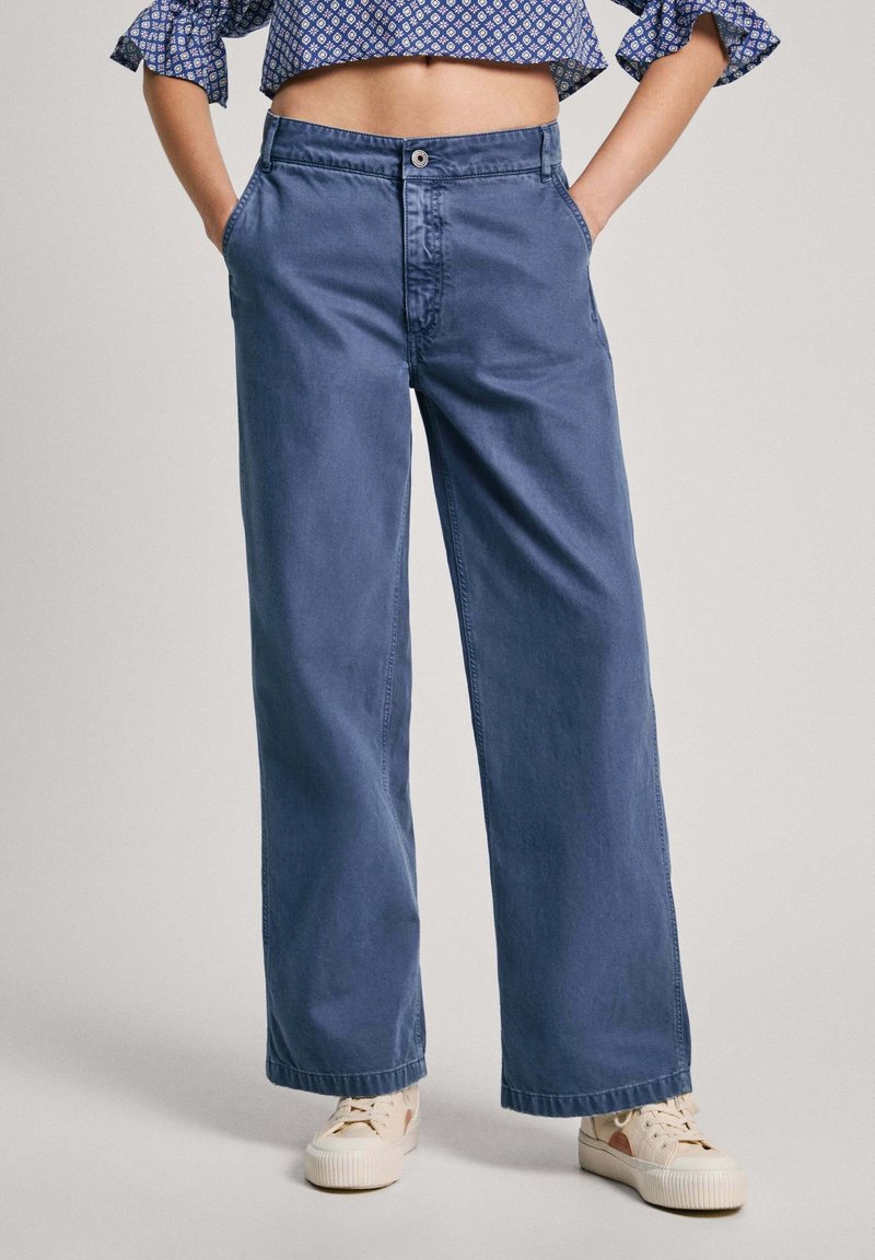 Pantalones vaqueros de pierna ancha en azul claro, con cierre de botones, dos bolsillos delanteros y un dobladillo sencillo. Combinados con zapatillas blancas.