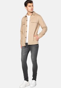 Beige jakke med knaplukning fortil, to brystlommer og lange ærmer, parret med mørkegrå jeans og hvide sneakers.
