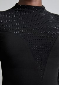 L�ähiotoksessa musta istuva vaate, jossa on pieniä heijastavia pisteitä järjestettynä geometriseen kuvioon rinnan ja hartioiden kohdalla.