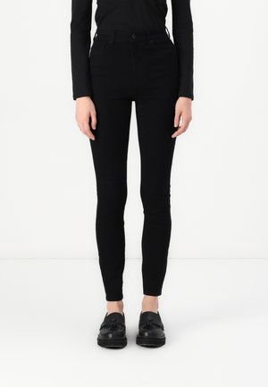 Person trägt eng anliegende schwarze Hose, schwarzen Langarm-Top und schwarze Tassel-Loafers, steht vor weißem Hintergrund.