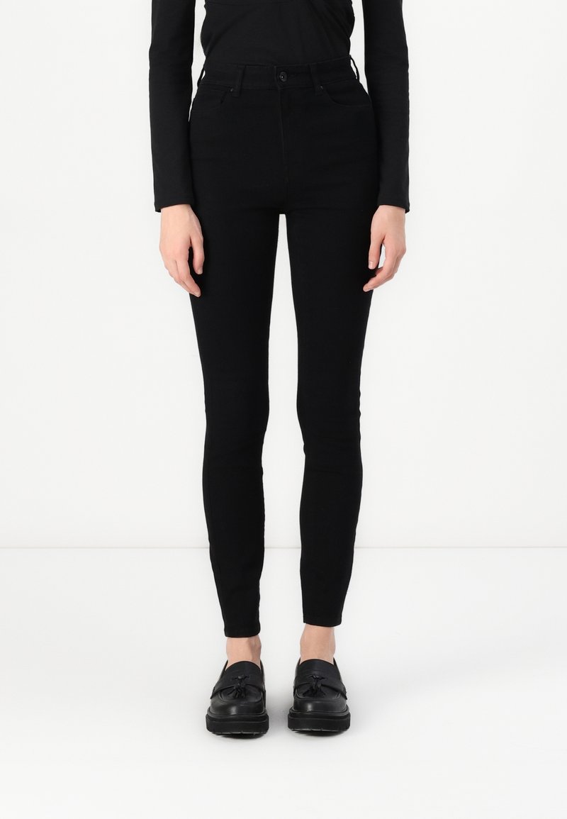 Personne portant un pantalon noir ajusté, un haut noir à manches longues et des mocassins noirs à pampilles, debout devant un fond blanc.
