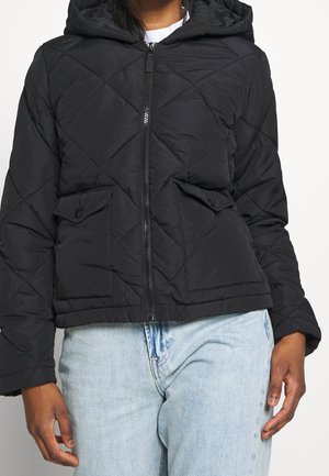 Person trägt eine schwarze gesteppte Kapuzenjacke mit Frontreißverschluss und Klappentaschen, kombiniert mit hellblauen Jeans aus Denim.
