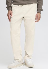 Beige cotton pants with a relaxed fit, featuring front pockets and a smooth texture, paired with light-colored athletic shoes.