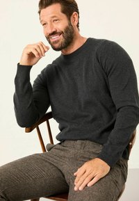 Dunkelgrauer Strickpullover mit gerippten Bündchen, kombiniert mit braunen Hahnentritthosen. Das Modell sitzt auf einem Holzstuhl, lächelt und hat die Hand an das Gesicht gelegt.