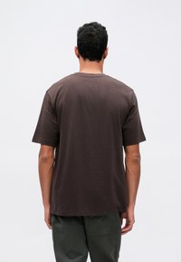 Lös passande brun bomullströja med rund halsringning och korta ärmar, visad bakifrån mot en enfärgad vit bakgrund.