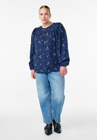 Mornarska bluzica s svetlo modrim cvetnim vzorcem, puffed rokavi in gumbastim ovratnikom, v kombinaciji z svetlo modrimi hlačami z visokim pasom in črnimi škornji.