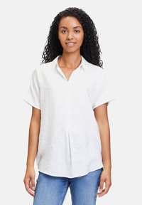 Weißes, kurzärmliges Hemd aus strukturiertem Stoff mit durchgehender Knopfleiste und lockerer Passform. Kombiniert mit hellblauen Jeans.