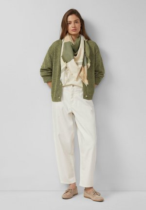 Frau steht vor einer grauen Wand, trägt einen grünen Cardigan, einen beigen und grünen Schal, weiße hoch taillierte Hose und beige Mokassins.