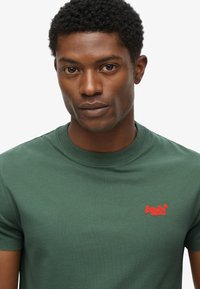 Mand med kort, krøllet hår iført en grøn SuperDry T-shirt med rødt logo på venstre bryst, der ser direkte frem mod en hvid baggrund.