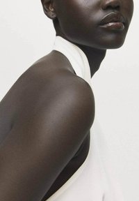 Un primer plano del hombro y el cuello de un modelo con una blusa de tela blanca, mostrando una piel oscura suave y un brillo labial natural contra un fondo claro.