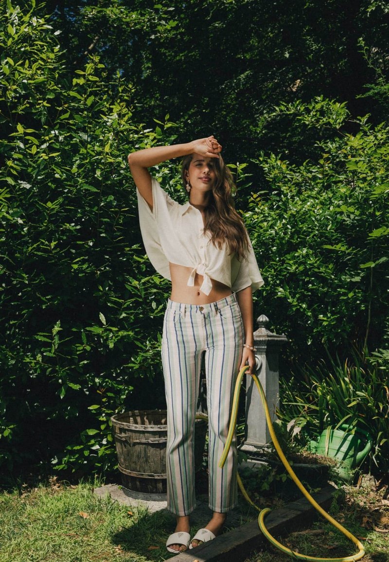 Jeune femme en pantalon rayé et chemise blanche nouée se tient dans le jardin, tenant un tuyau jaune, se protégeant les yeux de la lumière du soleil.