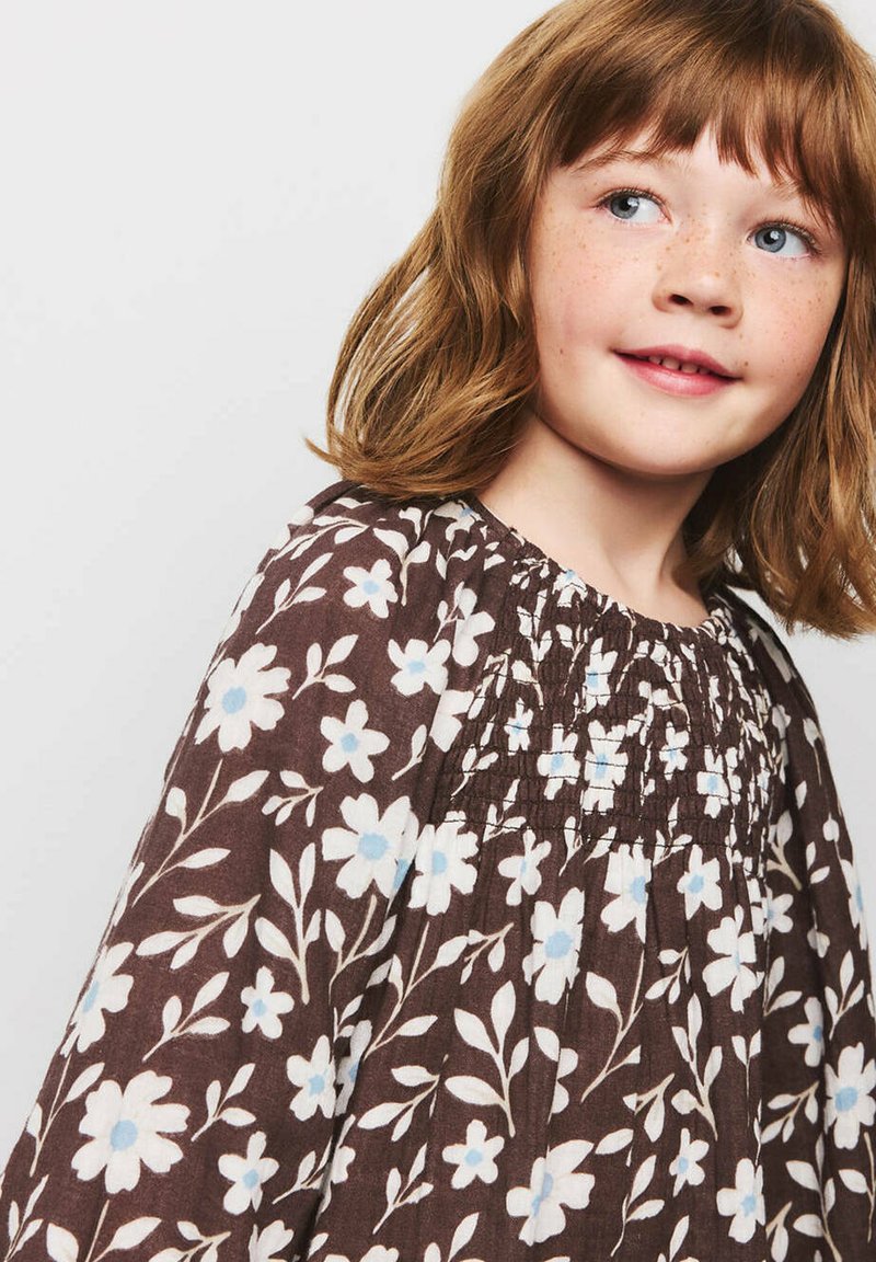 Jeune fille rousse avec des taches de rousseur portant une robe marron à motifs de fleurs blanches et bleu clair, regardant légèrement sur le côté.