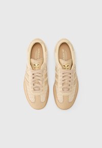 Sapatos de atletismo beige com parte superior em malha texturizada e couro liso, três riscas beige e um detalhe em logo dourado na língua.