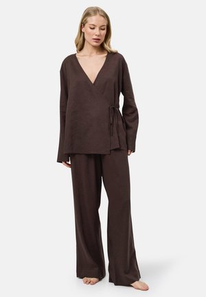 Blonde vrouw die een losse donkerbruine wikkeltop met zijstrik en wijde broek draagt, staat blootsvoets op een lichte achtergrond.