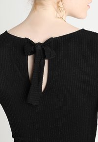 Vue de dos d'une femme portant une robe noire côtelée avec un nœud noué dans le haut du dos et des boucles d'oreilles géométriques dorées.