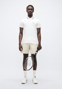 Λευκό πόλο πουκάμισο με μαύρες λεπτομέρειες στον γιακά, συνδυασμένο με μπεζ σορτς. Το μοντέλο κρατά ρακέτα τένις, φορώντας λευκές κάλτσες και παπούτσια.