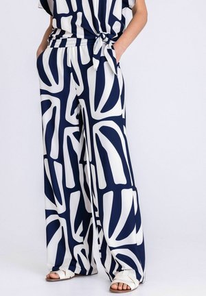 Vrouw draagt losse wijde broek met marineblauw-wit patroon, handen in de zakken en witte sandalen op witte achtergrond.