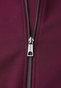 Bordeaux zip-up plagg med en metallisk dragkedja, texturerad tygfinish och en logotyp på dragkedjans pull.