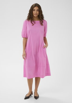 Žena s kučeravými vlasmi v ružových midi šatách s nafúknutými rukávmi a čiernymi nízkymi sandálmi na opätku, stojaca s jednou rukou v vrecku.