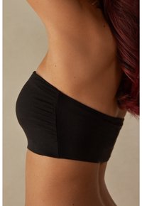 Mustat bandeau-top, jossa on sileä pinta ja rypytetty muotoilu sivuilla, korostaen saumattoman istuvuuden neutraalilla taustalla.