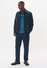 Jachetă din bumbac bleumarin cu închidere prin buton, asociată cu o cămașă din denim și pantaloni asortați. Are o textură netedă și un design minimalist.