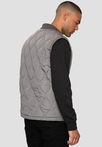 Grau gesteppte Weste mit Stehkragen, die über einem schwarzen Langarmshirt getragen wird. Sie zeichnet sich durch eine glatte Textur und abgerundete Steppnähte aus.