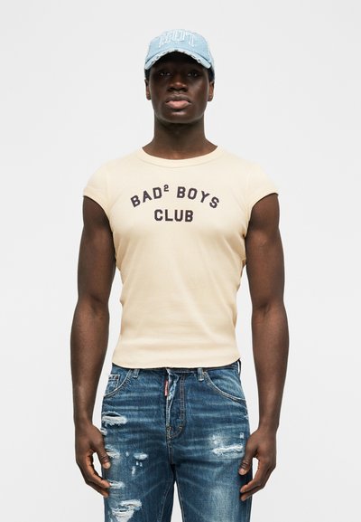 Άνδρας με μπεζ αμάνικη μπλούζα με το κείμενο "BAD² BOYS CLUB", φθαρμένο μπλε τζιν και ανοιχτό μπλε καπέλο, στέκεται μπροστά σε λευκό φόντο.