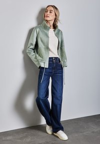 Giacca in eco-pelle verde con dettagli in pelliccia, abbinata a jeans a gamba larga blu scuro e sneakers bianche su uno sfondo neutro.