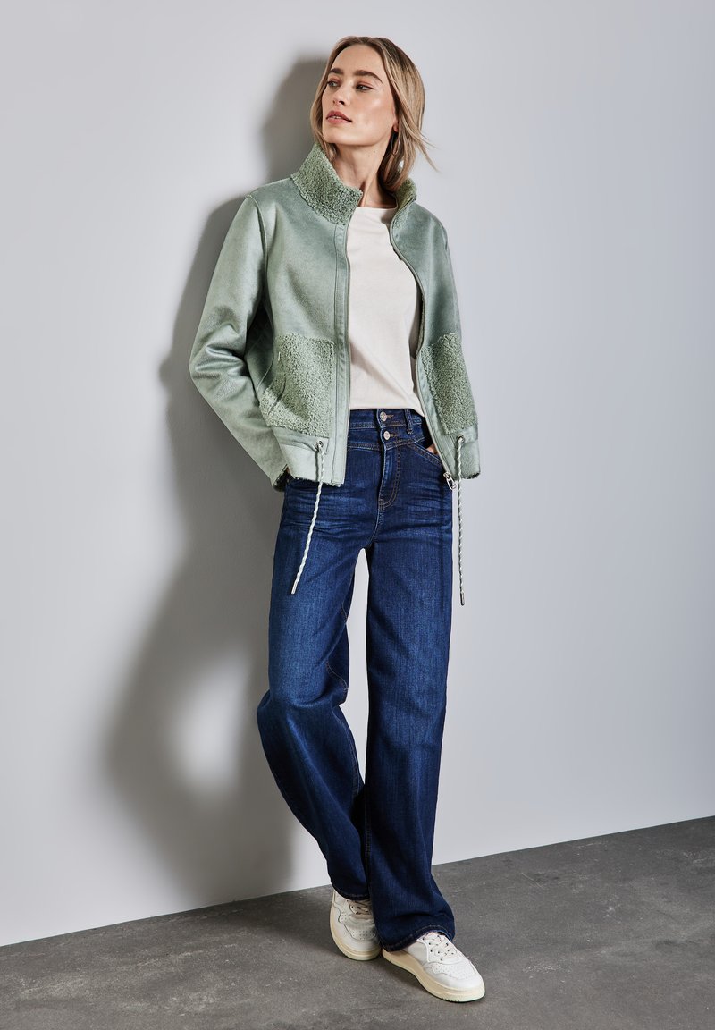 Giacca in eco-pelle verde con dettagli in pelliccia, abbinata a jeans a gamba larga blu scuro e sneakers bianche su uno sfondo neutro.