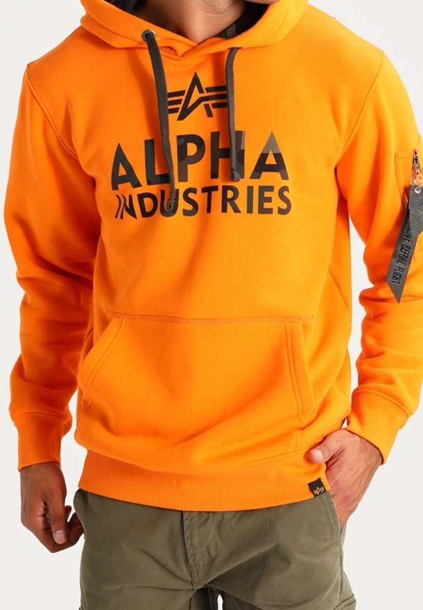 Homme portant un sweat à capuche Alpha Industries orange vif avec logo noir, cordons, poche avant et pantalon vert, main dans la poche.