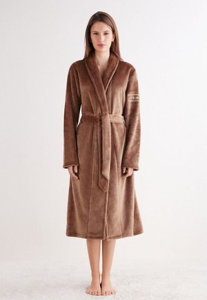 Frau steht barfuß in einem langen, braunen Plüschbademantel, der in der Taille gebunden ist, vor einem schlichten weißen Hintergrund.