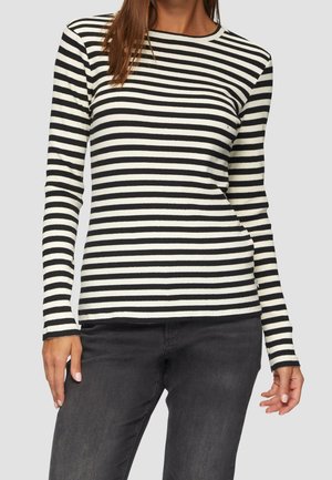 Femme portant un t-shirt à manches longues rayé horizontalement noir et blanc et un jean gris foncé, debout devant un fond uni.