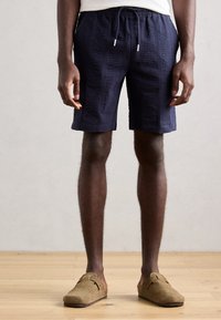 Marineblauwe shorts met een gestructureerd patroon, een elastische tailleband met trekkoord en een relaxte pasvorm, gecombineerd met bruine instappers.
