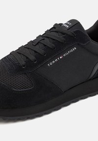 Fekete sneaker, amely nubuk és hálós anyagok keverékéből készült, párnázott talppal. Az oldalán "TOMMY HILFIGER" felirattal. Fűzők a biztos illeszkedés érdekében.
