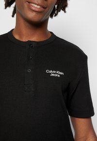 Černé tričko s krátkým rukávem typu henley s žebrovanou texturou, které má tři knoflíky a bílé logo "Calvin Klein Jeans" na přední straně.