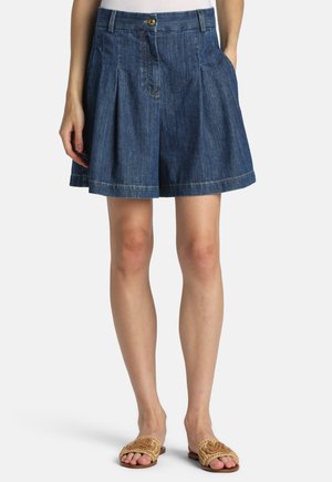 Denim plisserede shorts med knap- og lynlåslukning båret sammen med vævede slip-on sandaler på model mod hvid baggrund.
