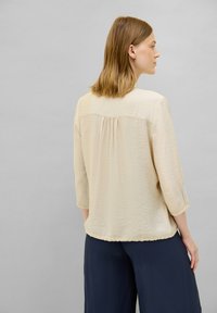 Beige, lichtgewicht blouse met een textuurafwerking, 3/4 mouwen en een relaxte pasvorm. De achterkant heeft een plooidetail voor extra accent. Gemaakt in combinatie met donkere broeken.
